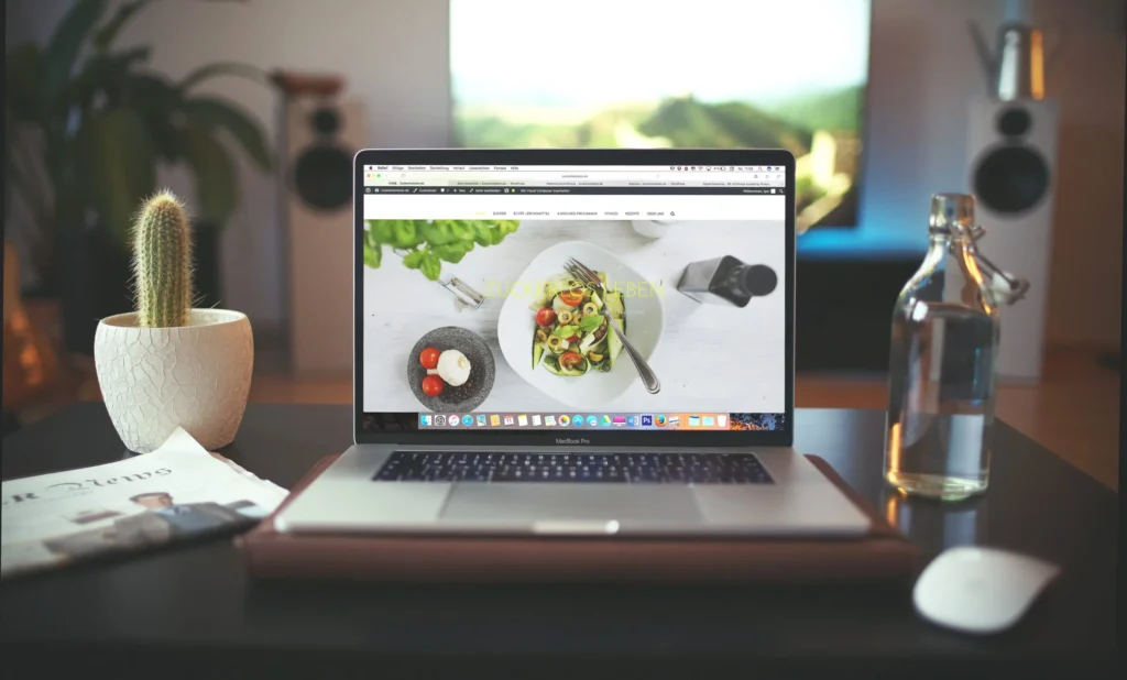 MacBook Pro displaying zuckerlosleben.de website with healthy food recipe, featuring water bottle, cactus, and newspaper on a modern desk setup.