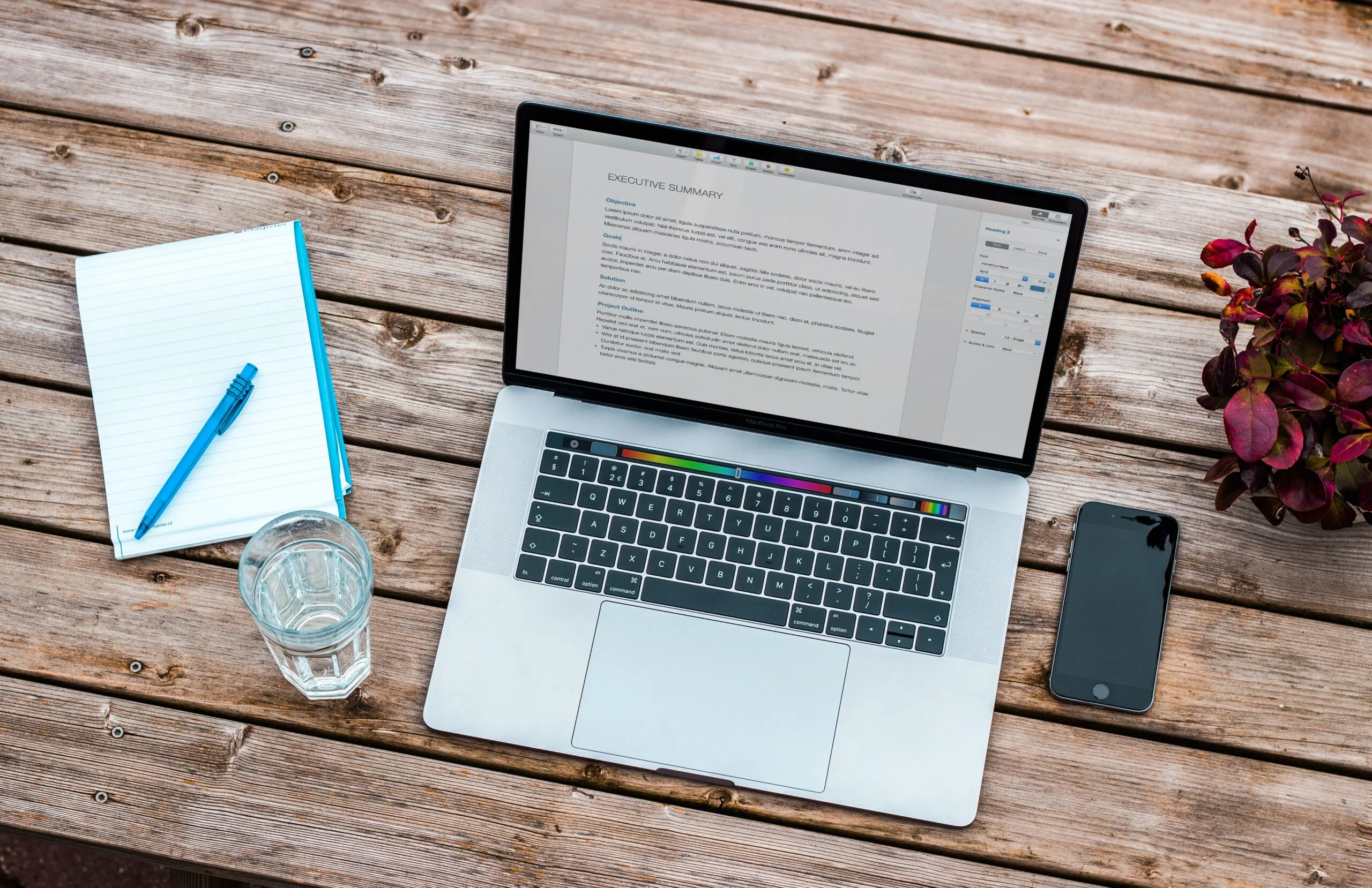 Open laptop on wooden table displays a document. Nearby are a notebook with a pen, a glass of water, a smartphone, and a potted plant, creating a calm workspace.