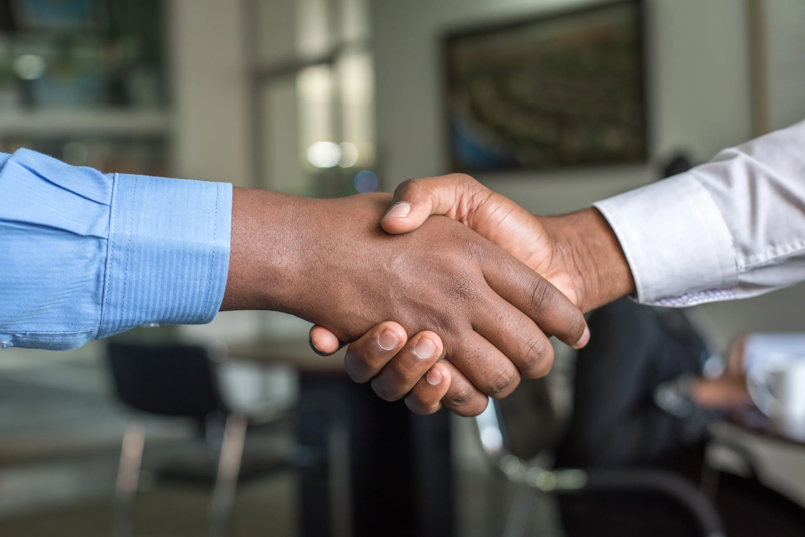 Two individuals are shaking hands in an office setting, symbolizing agreement or partnership. The atmosphere appears professional and cooperative.