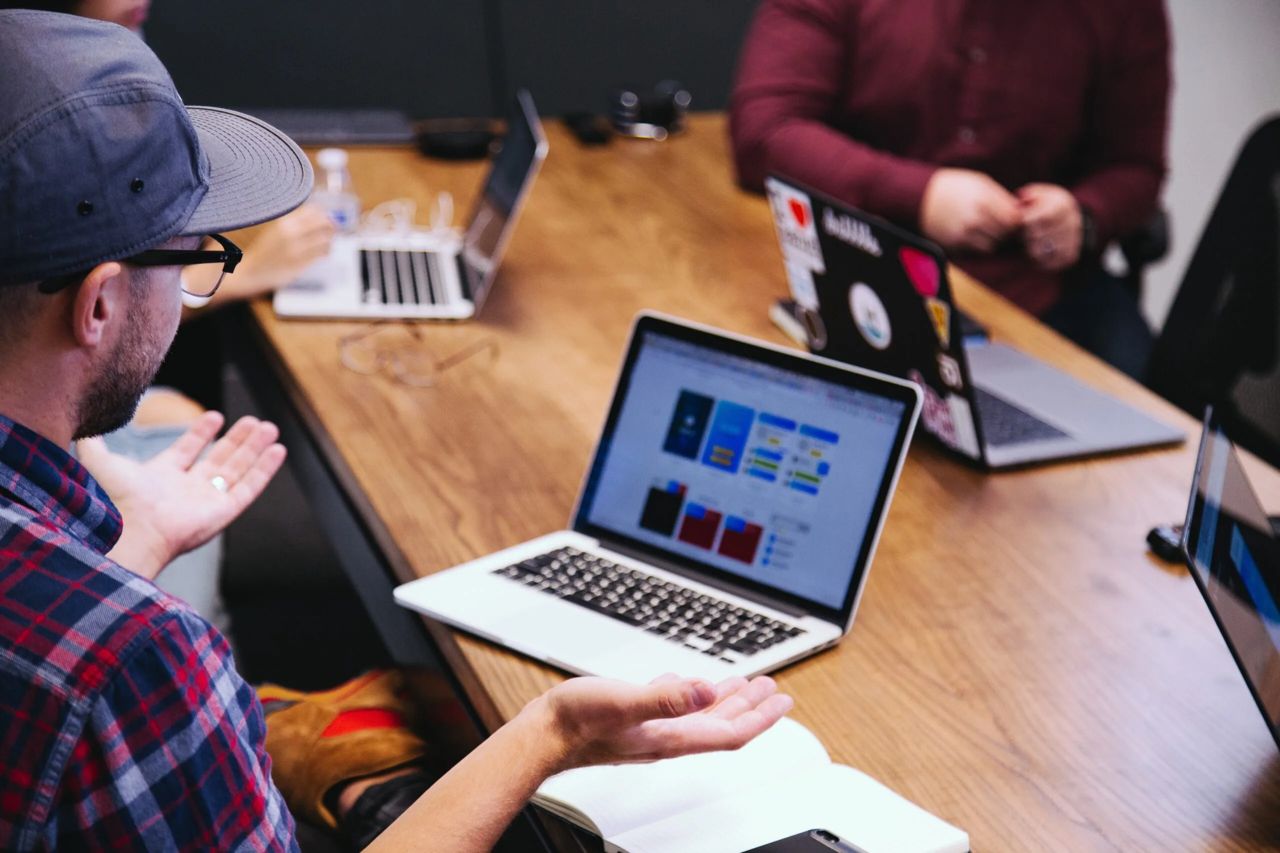 A group of people in a meeting room sitting around a wooden table with laptops open. One person gestures while discussing a colorful laptop screen. The atmosphere is collaborative.