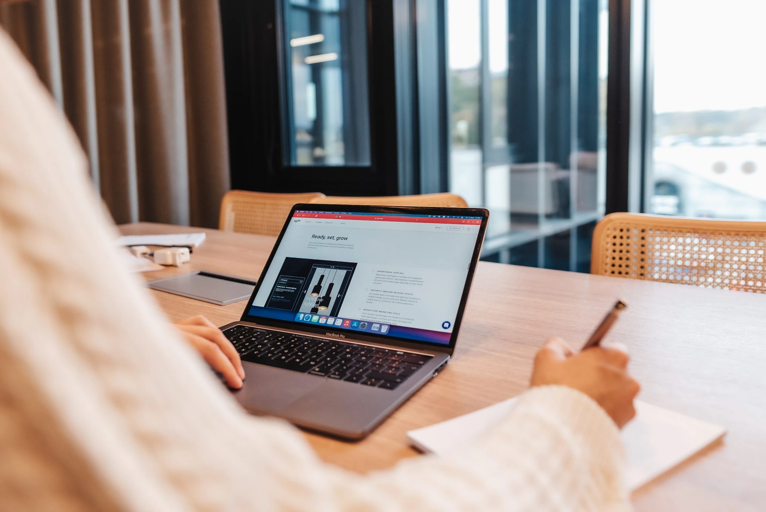 A person is sitting at a wooden table, working on a MacBook Pro while taking notes with a pen on a white notepad. The laptop screen displays a webpage titled "Ready, set, grow," with images of lighting fixtures. A trackpad rests beside the laptop. The setting appears to be an office or co-working space with large windows, allowing natural light to illuminate the workspace. The person's arm and part of their torso are visible, and they are wearing a cream-colored sweater. Wicker chairs are partially visible behind the table.